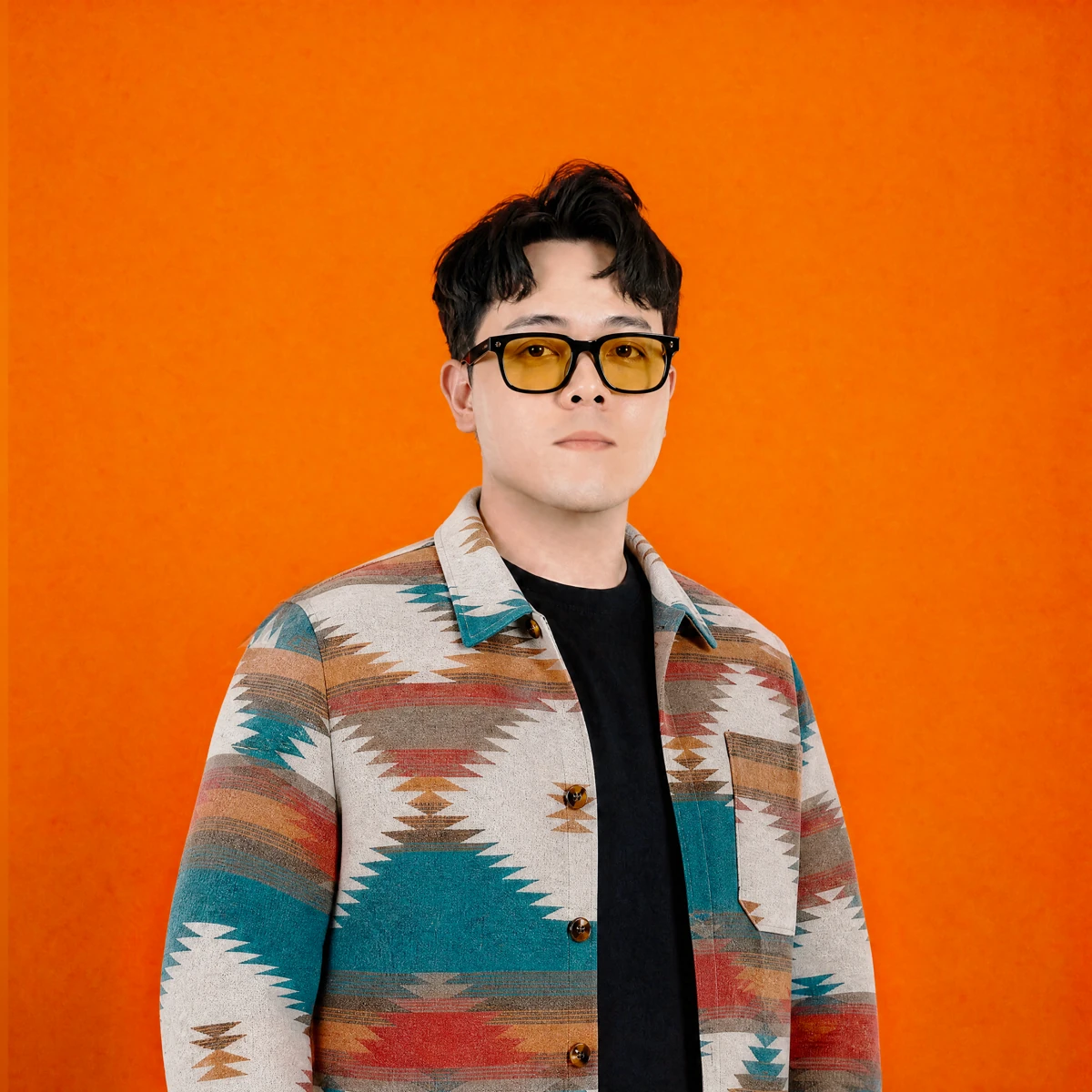 Syahkeer Rodrigues wearing amber glasses and a patterned jacket against an orange studio backdrop
